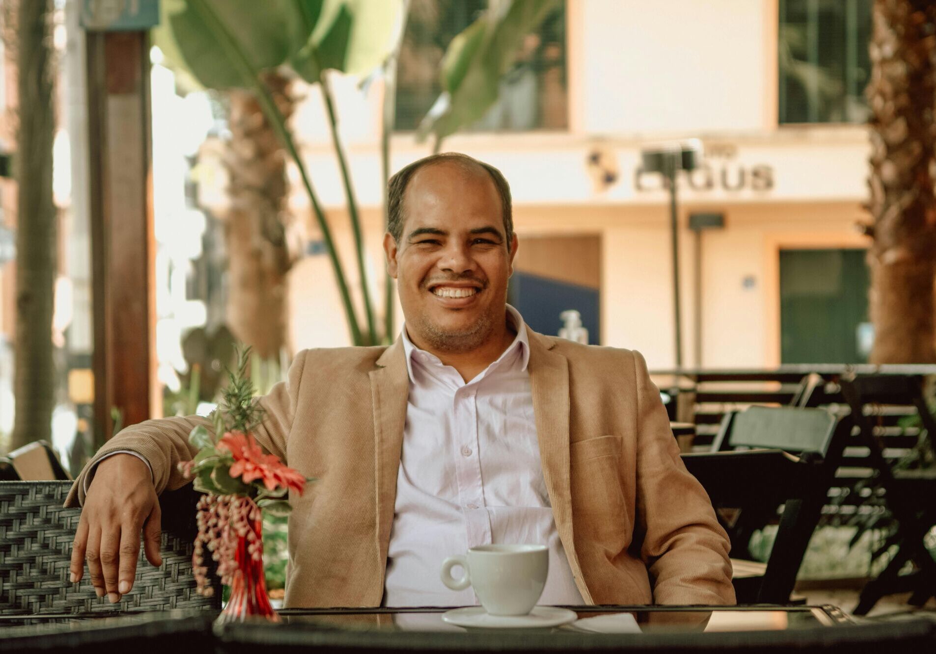 Balding man in a suit jacket smiling while enjoying a coffee in an outdoor café setting.